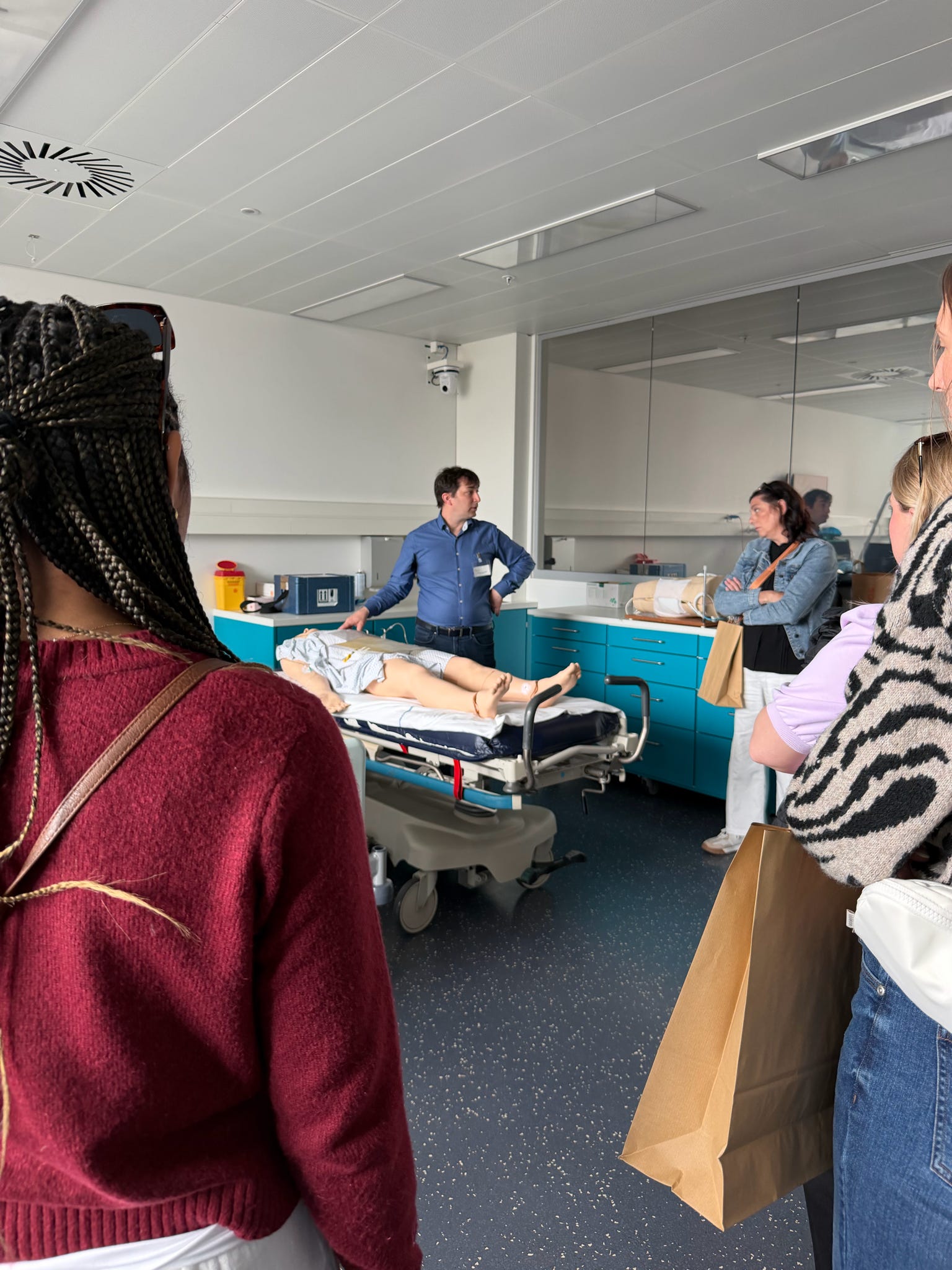 gather around a hospital bed while an instructor demonstrates a medical procedure on a training mannequin.