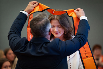 A person places an orange honors stole around another person’s shoulders during an indoor recognition ceremony, marking an academic award or distinction.