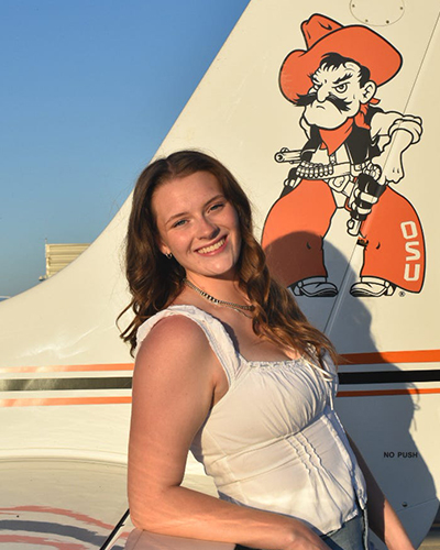 A person poses outdoors beside an aircraft tail decorated with an illustrated OSU Cowboy mascot, with the airplane’s white and orange paint visible against a clear sky, suggesting a campus or aviation-themed photo setting.