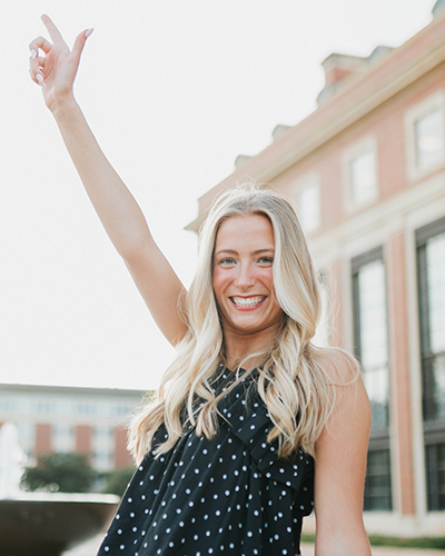 A person stands outdoors on a college campus with one arm raised, wearing a sleeveless dark top with a dotted pattern, posed in front of academic buildings in a bright, sunlit setting.