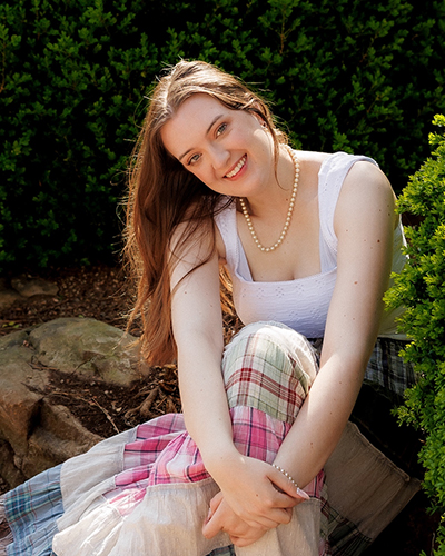 A person sits outdoors among rocks and green shrubs, wearing a light-colored sleeveless top and patterned pants, posed for a casual portrait in a landscaped garden setting.