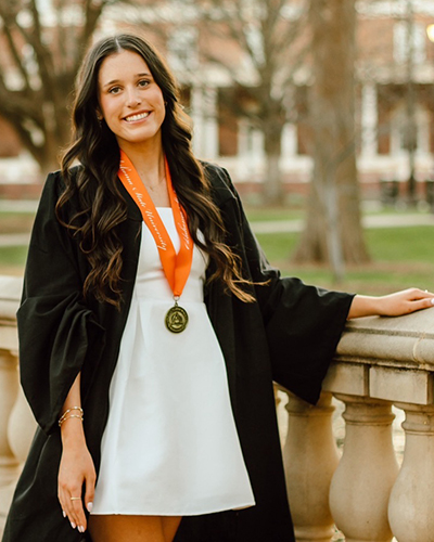 A person in a graduation gown stands outdoors by a stone railing on a campus walkway, wearing an academic medal on an orange ribbon over a light-colored dress with trees and buildings in the background, indicating a graduation portrait setting