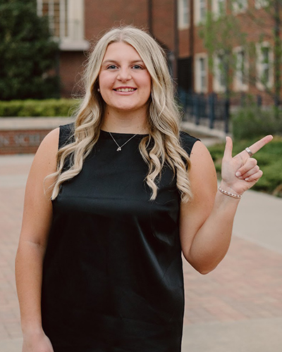 A person stands on a brick campus walkway wearing a sleeveless black top and pointing to the right, with campus landscaping and academic buildings visible in the background, suggesting a posed campus portrait.