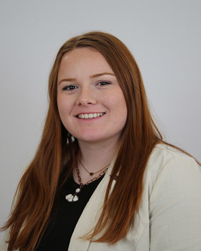 A professional studio headshot shows a person with long hair wearing a light-colored blazer over a dark top, facing the camera against a plain neutral background.