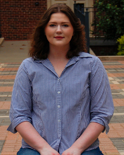 A person stands on a campus brick walkway wearing a blue-and-white striped button-down shirt, posed facing the camera with academic buildings and walkways visible in the background.