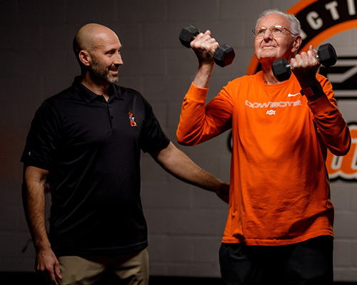 One person supervises as another person lifts two dumbbells in an indoor training facility, demonstrating a weightlifting exercise in front of an OSU-branded backdrop.