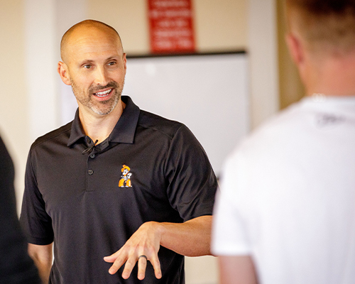 A person wearing an OSU-branded black polo gestures while speaking to another individual in an indoor training or classroom setting, with a whiteboard visible in the background.