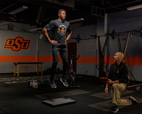 In an indoor gym, an athlete performs a box jump onto a raised platform while a trainer kneels nearby holding a tablet and monitoring the exercise, with OSU branding and workout equipment visible in the background.