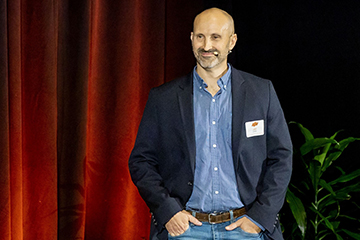 A person wearing a blazer and jeans stands on a stage with a red curtain backdrop, speaking during a presentation while a potted plant sits nearby.