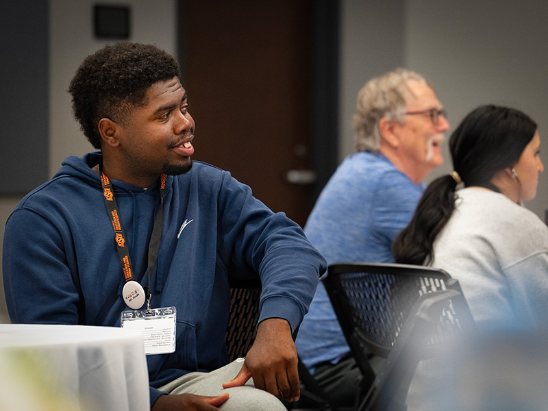 A group of attendees sits at tables during an indoor program or workshop, with one person in the foreground wearing a name badge and others listening and engaging in discussion in the background.