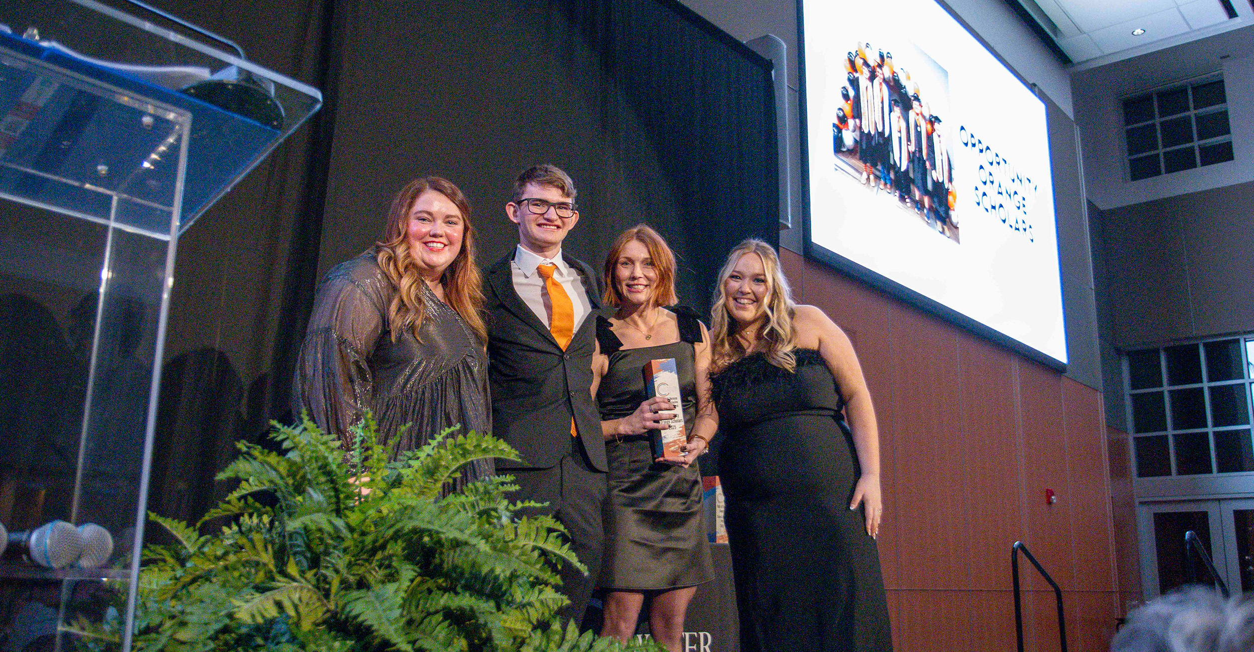 Four people stand together on a stage holding an award in front of a large screen and podium, documenting a formal recognition ceremony at a university or community awards event.