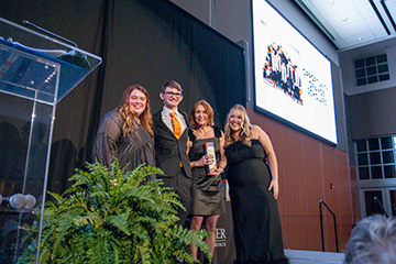 Four people stand together on a stage holding an award in front of a large screen and podium, documenting a formal recognition ceremony at a university or community awards event.