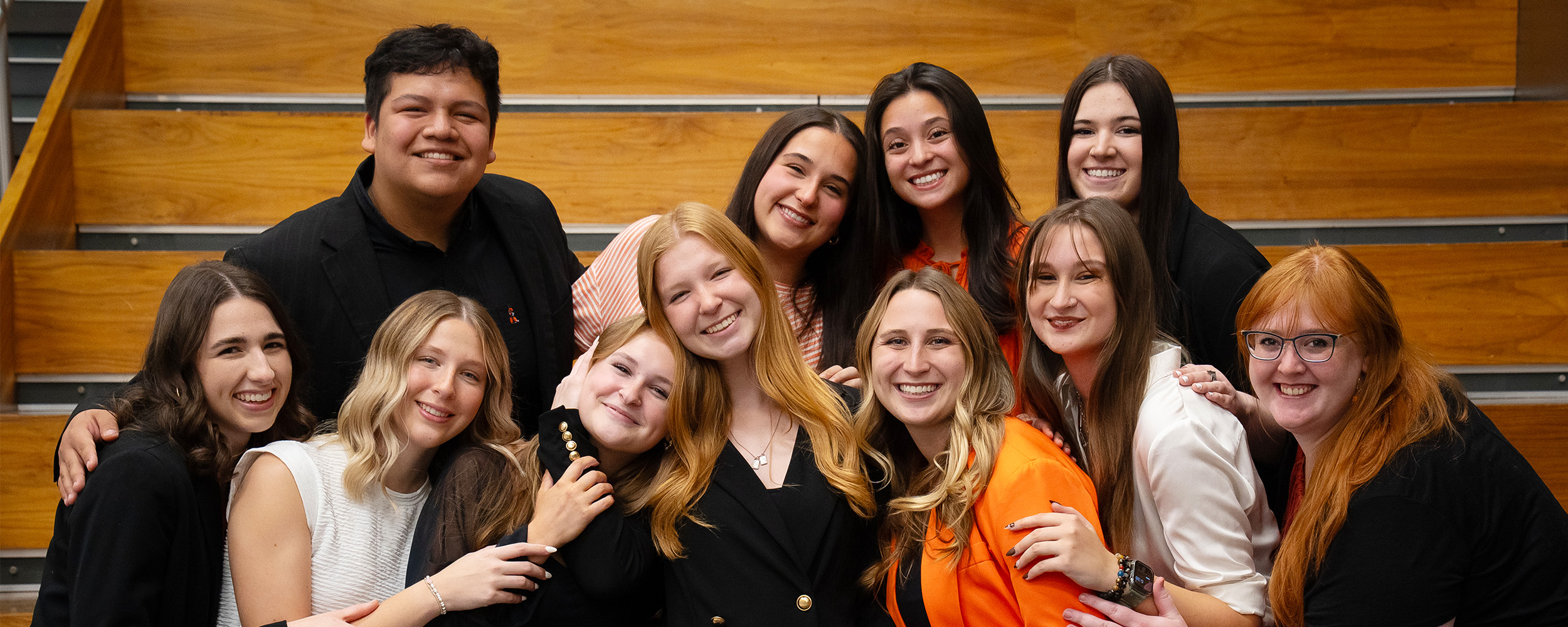 A group of 11 people sit and stand closely together on indoor wooden bleachers, posing as a team portrait while dressed in a mix of business attire and casual clothing.