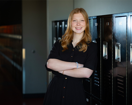 A person with long hair wearing a black dress stands with arms crossed in a school hallway, posed in front of a row of dark lockers for a portrait-style photo.