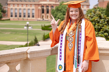 A graduate in an orange cap and gown with multiple honor cords stands on a stone terrace in front of a campus building, raising one finger while posing for a celebratory graduation portrait.