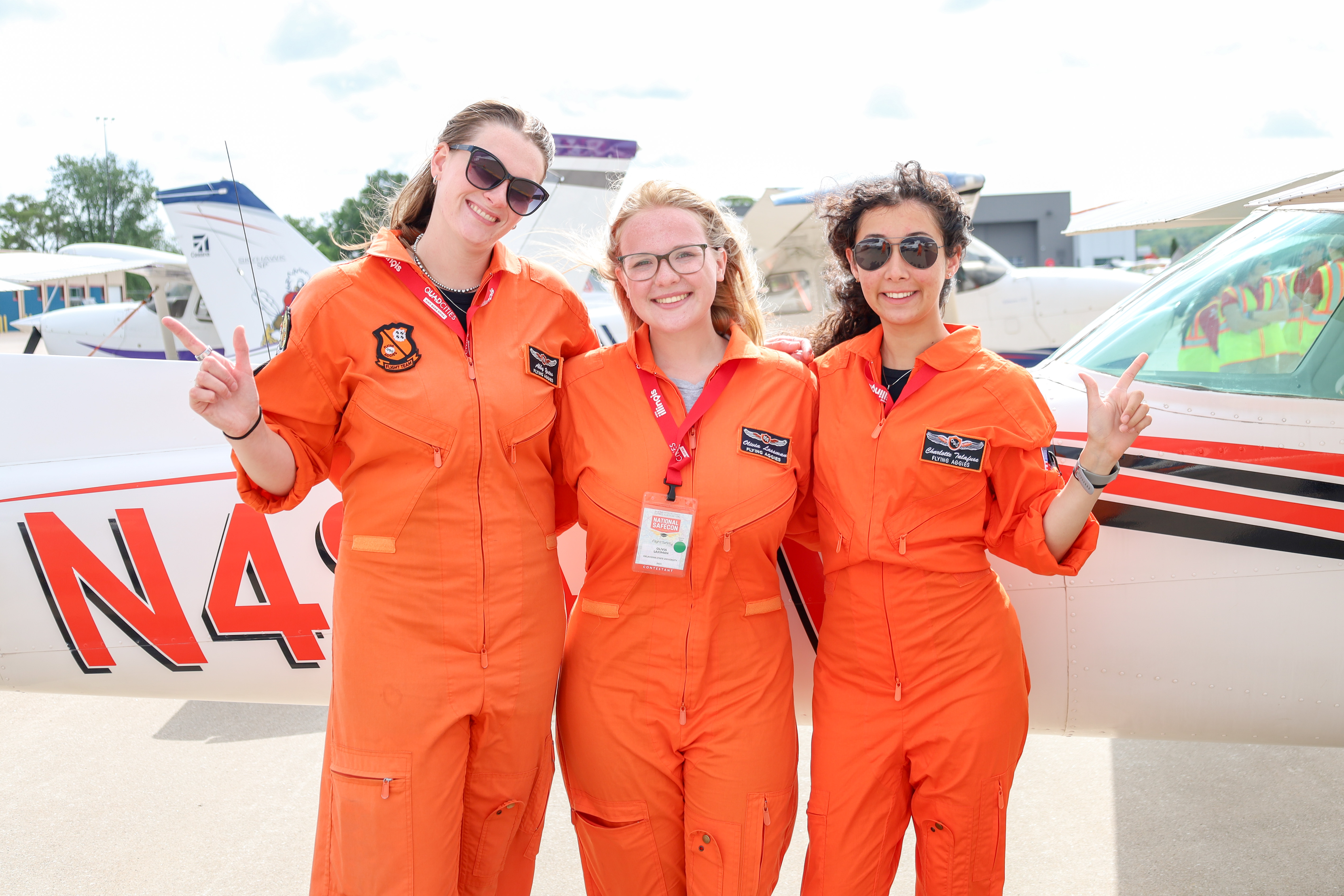 Three women in orange flight suits stand together posing in front of airplanes with 