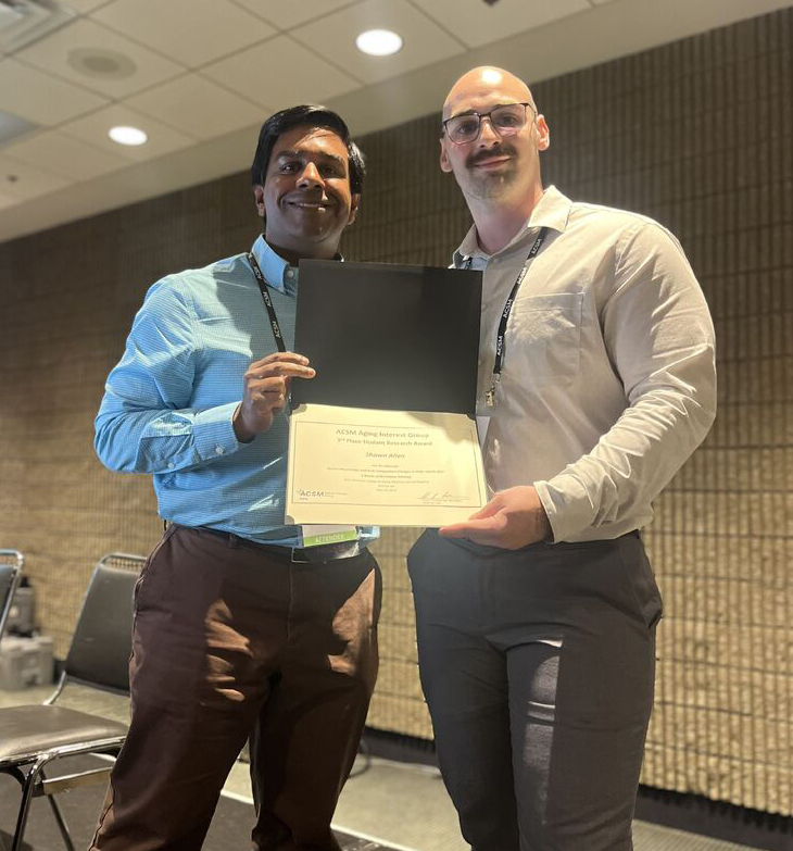 Two people standing side by side holding a certificate together, one wearing a blue dress shirt and brown pants, the other in a light-colored dress shirt and gray pants, in an indoor setting with chairs and a textured wall in the background.
