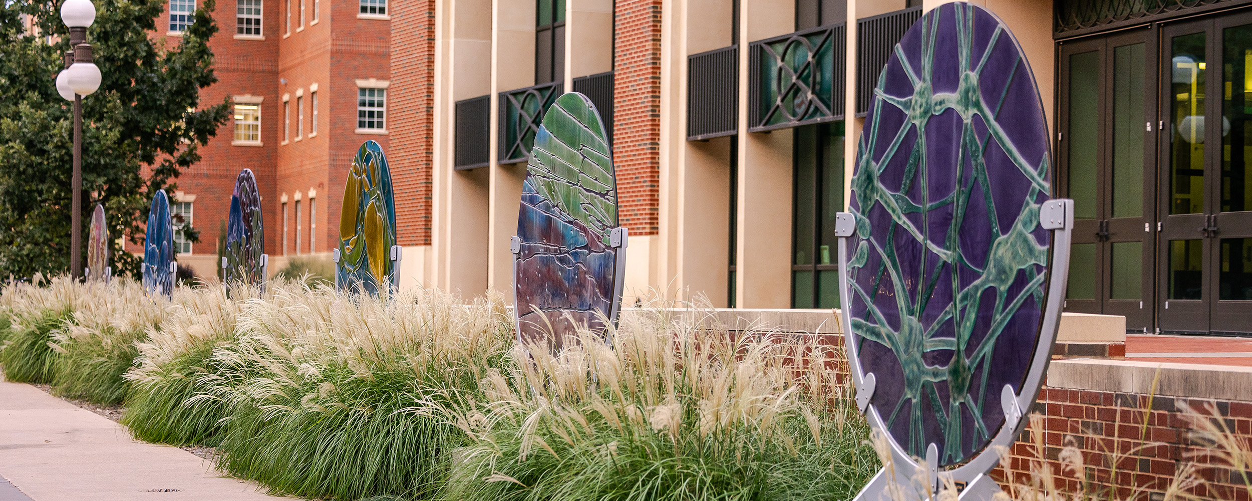 A row of large, colorful circular art pieces with abstract biological designs lines a landscaped walkway in front of a modern brick and glass building.