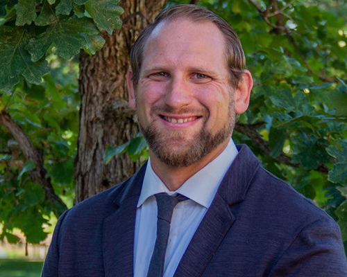 Color headshot of a person in a suit and tie standing outdoors in front of a tree with green leaves.
