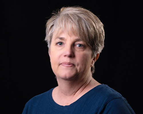 Color studio-style headshot of a person with short gray hair, wearing a teal shirt, facing the camera against a black background.
