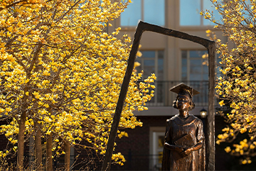 Color photograph of a bronze graduate statue framed by a rectangular metal structure, surrounded by blooming yellow trees with a campus building in the background.