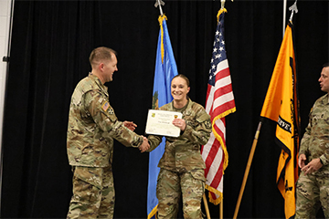 Clara receives a certificate during a formal military recognition ceremony, standing before three flags as two others look on.