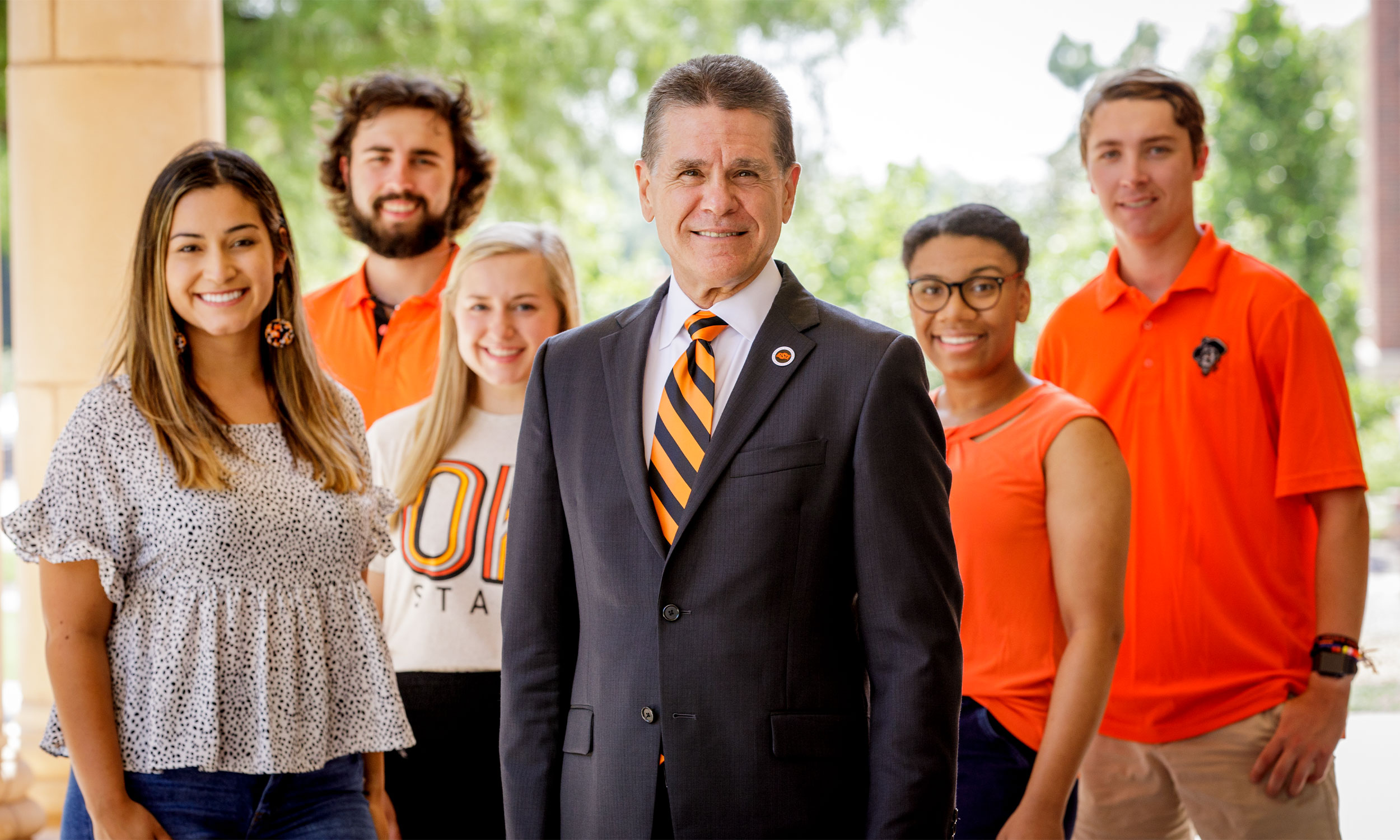 Group of six people standing outdoors with the central figure wearing a dark suit and an orange and black striped tie while the others wear casual orange and white outfits, with greenery and part of a building structure in the background.
