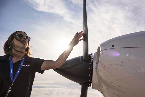 Woman in aviator glasses to the left of an air plane with her right hand trailing across the top of the propeller.