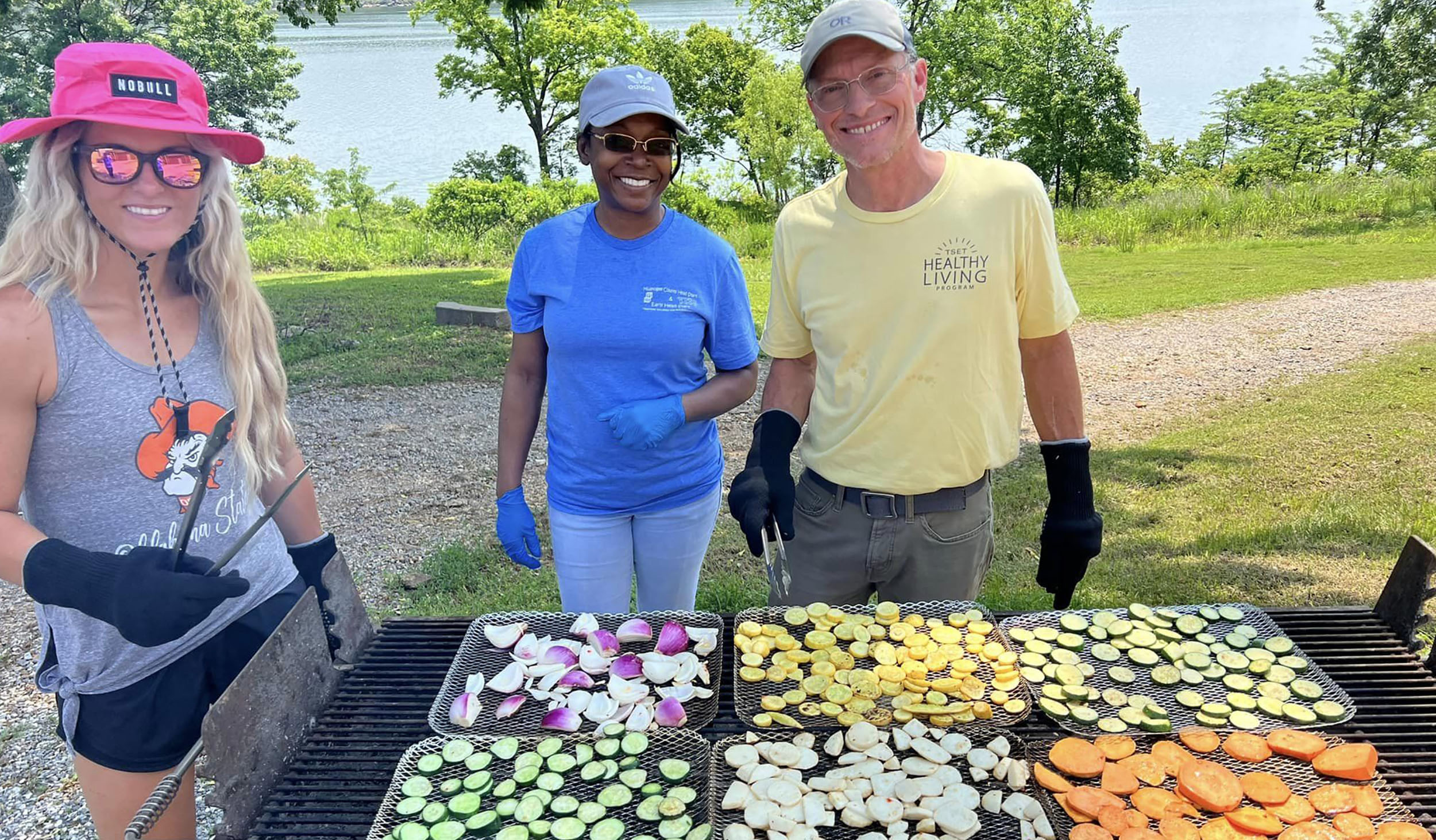three individuals stand over a grill cooking vegetables