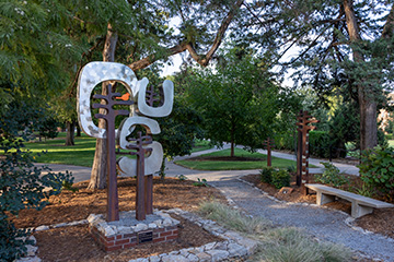 A metal abstract sculpture stands beside a paved walkway and wooden bench in a tree-lined outdoor garden with landscaped greenery