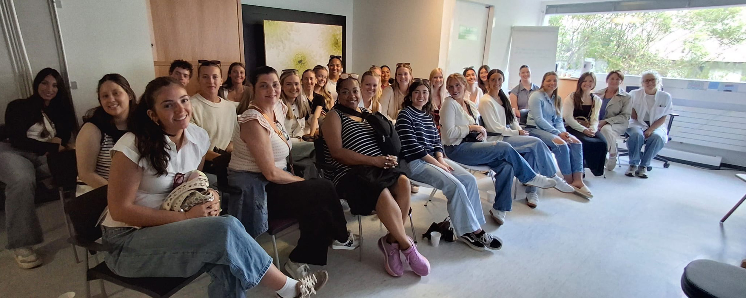 A group of people sit in rows in a bright classroom, attentively participating in what appears to be a lecture or training session.
