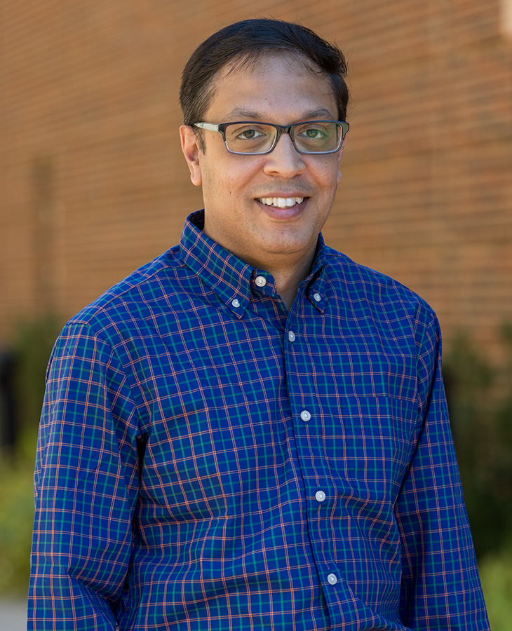 A man wearing glasses and a blue plaid shirt stands in front of a brick wall, smiling.