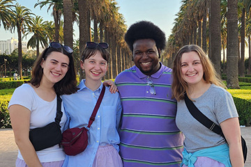 a group of students stand in front of a long path lined with palm trees all smiling, all wearing purple
