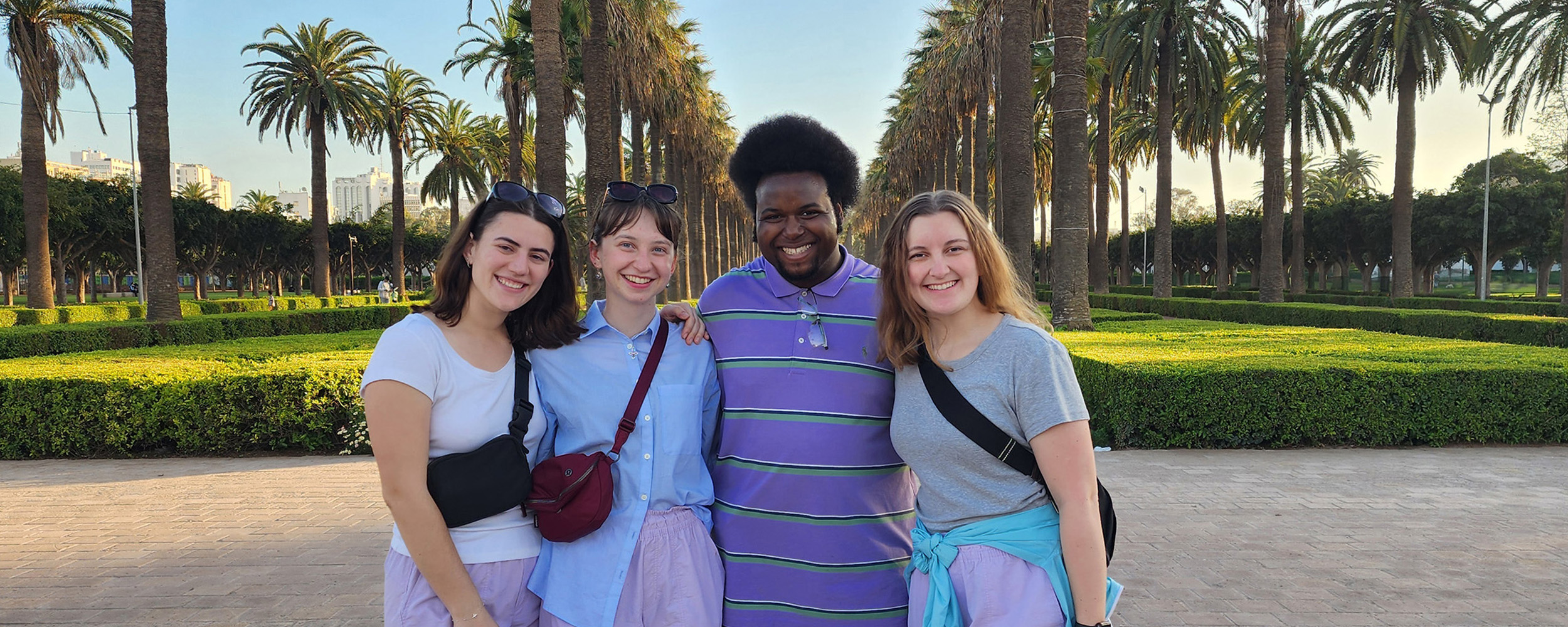 a group of students stand in front of a long path lined with palm trees all smiling, all wearing purple