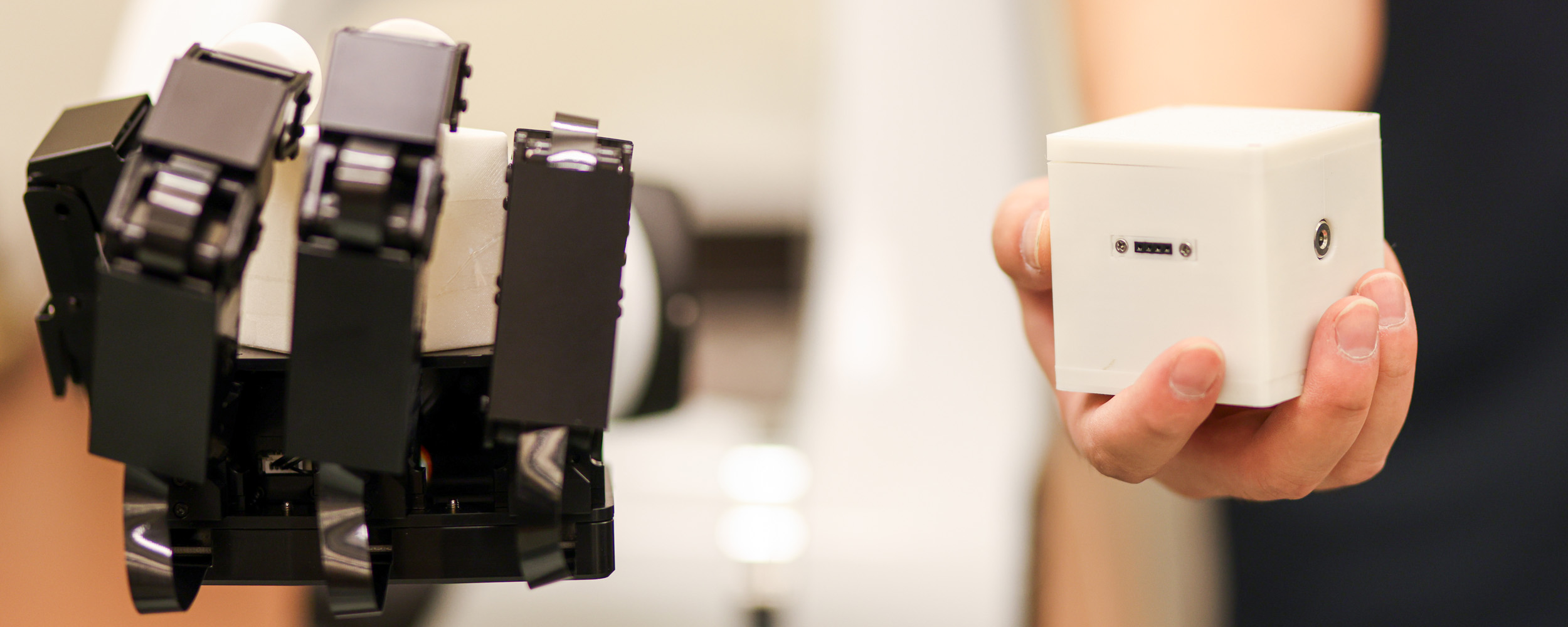 A robot hand beside Dr. Tao's hand holding a white cube that allows the robot hand to mimic the human motions.
