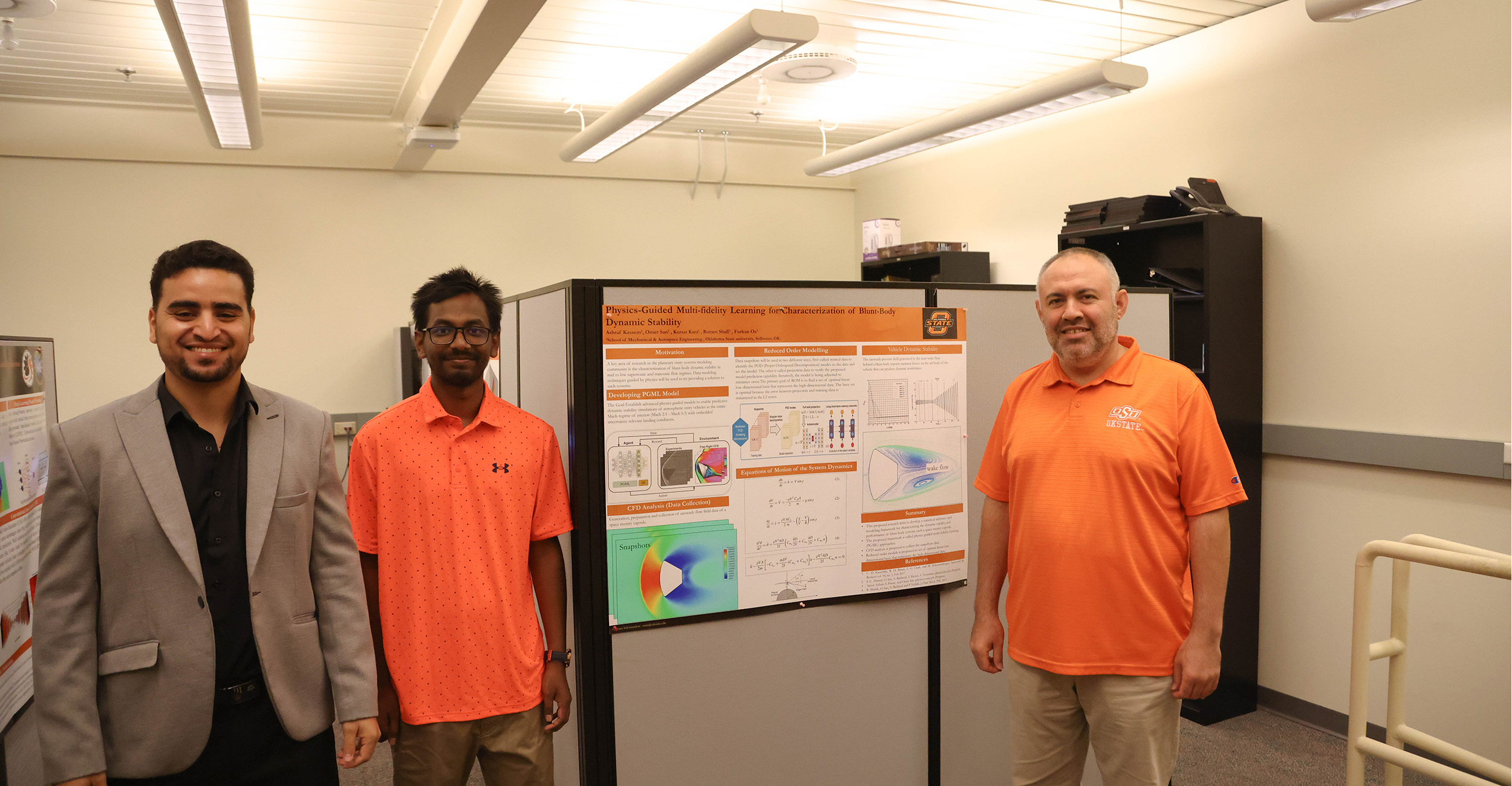 The image shows three men standing in a well-lit room with a white ceiling. They are all smiling and appear to be presenting a research poster titled "Physics Guided Multi-fidelity Learning for Characterization of Blm-Body Dynamic Stability". Two of the men are dressed casually in polo shirts, one orange and one coral pink