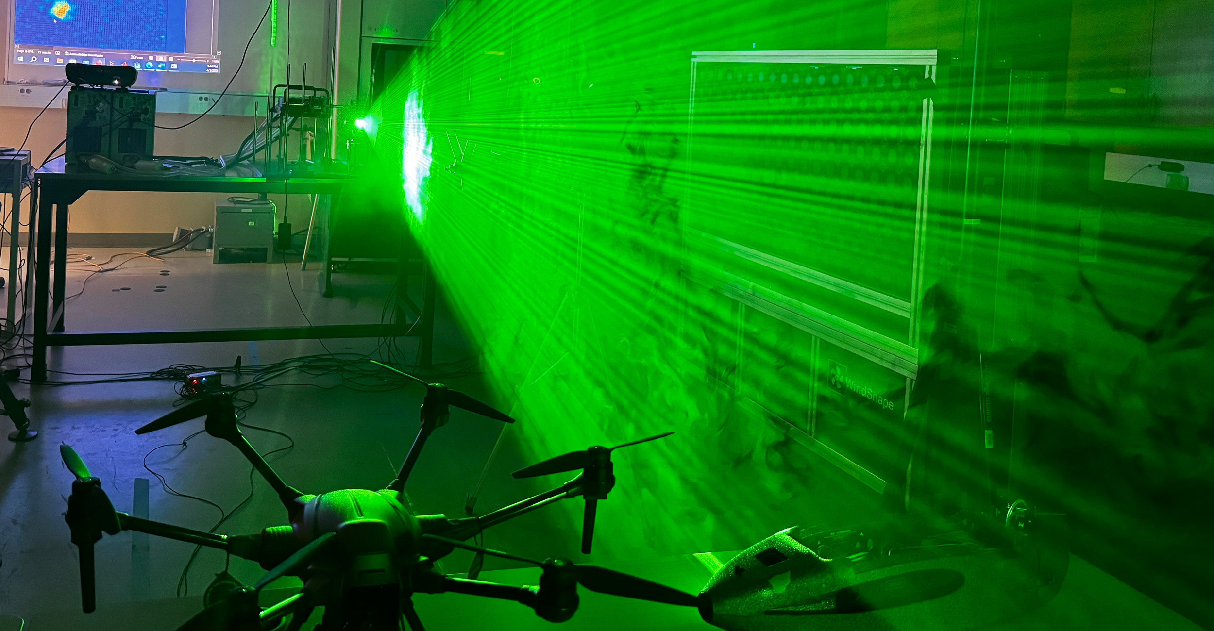 A green laser light fills a lab to show air movement with a large drone in the bottom left corner in a projector screen in the background with a graph of data.