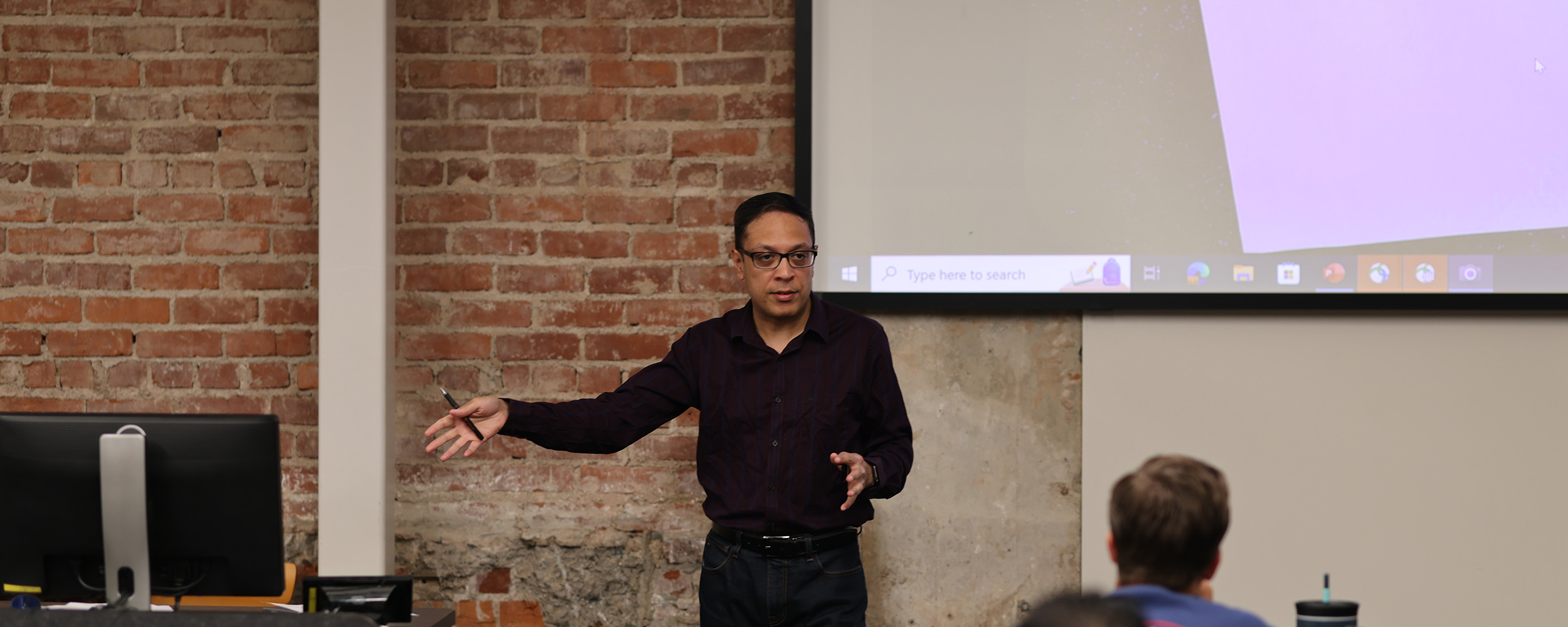 A man stands in front of a projector screen and brick wall, gesturing with his hands, while addressing an audience seated in the foreground.