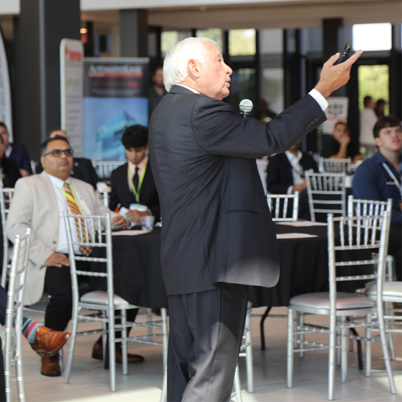 A man presenting at the conference holds the mic while gesturing towards the screen.