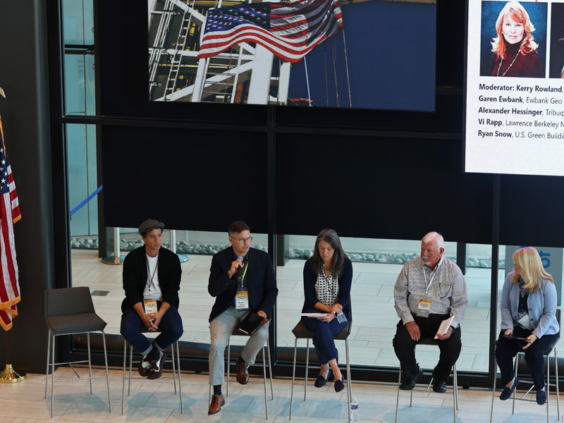 A panel of speakers sit in chairs at the front of a large room.