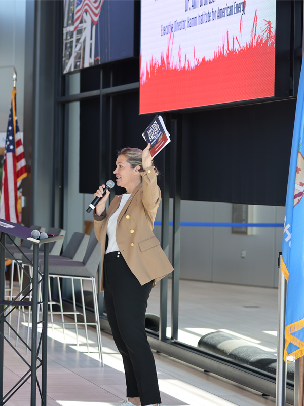 A woman speaker holds a mic infront of a large room of people while holding up a book to show the crowd in her other hand.
