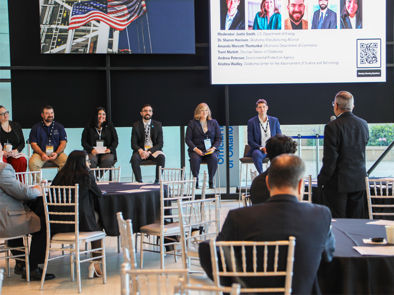 A panel of speakers sit in chairs at the front of a large room listening to a man from the crowd stand and ask a question into the mic.