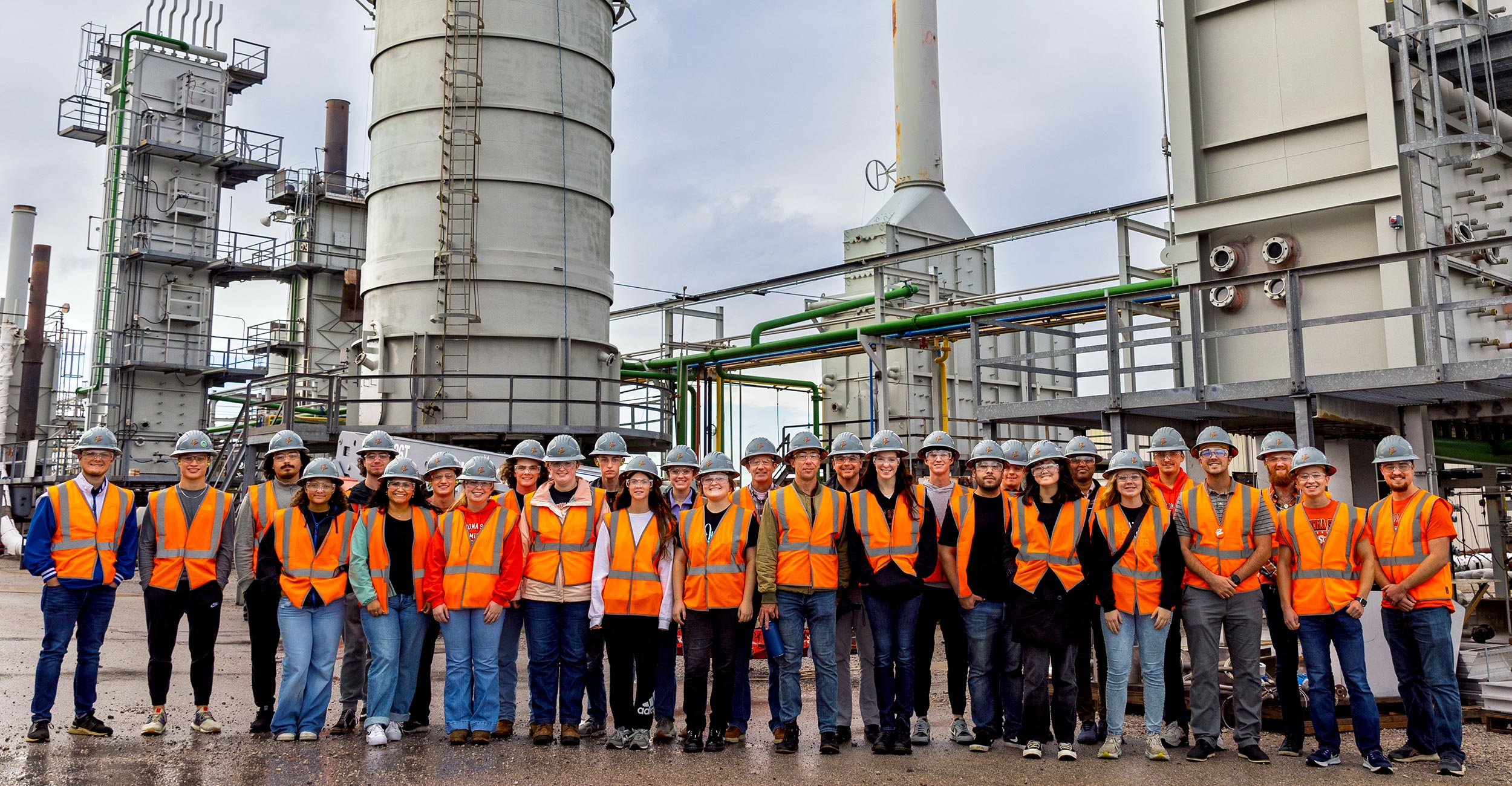 Smiling chemical engineering students standing in front of industrial buildings and equipment in orange vests and orange hardhats.