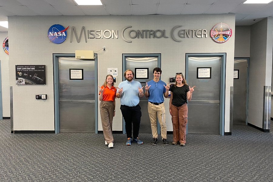 Four people pose underneath a sign that reads Mission Control Center at NASA's Johnson Space Center. 