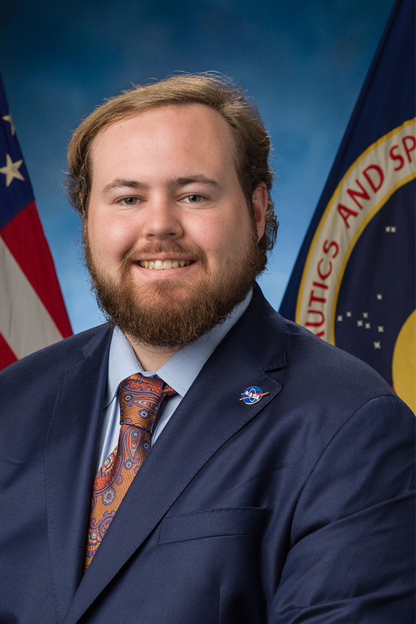 A well-dressed man with a beard stands before American flags, embodying a sense of patriotism and formal attire.