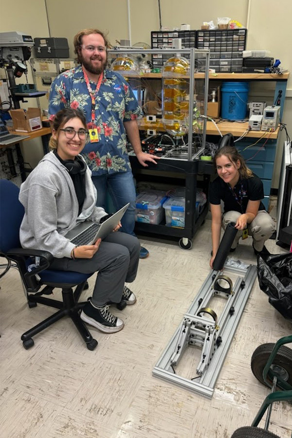 Three NASA interns are shown in a lab setting surrounded by equipment. Their smiles indicate a collaborative environment.