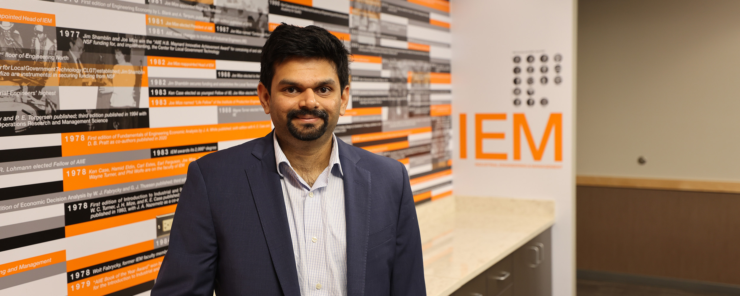 A man in formal attire stands before a wall marked with the letters "iem."