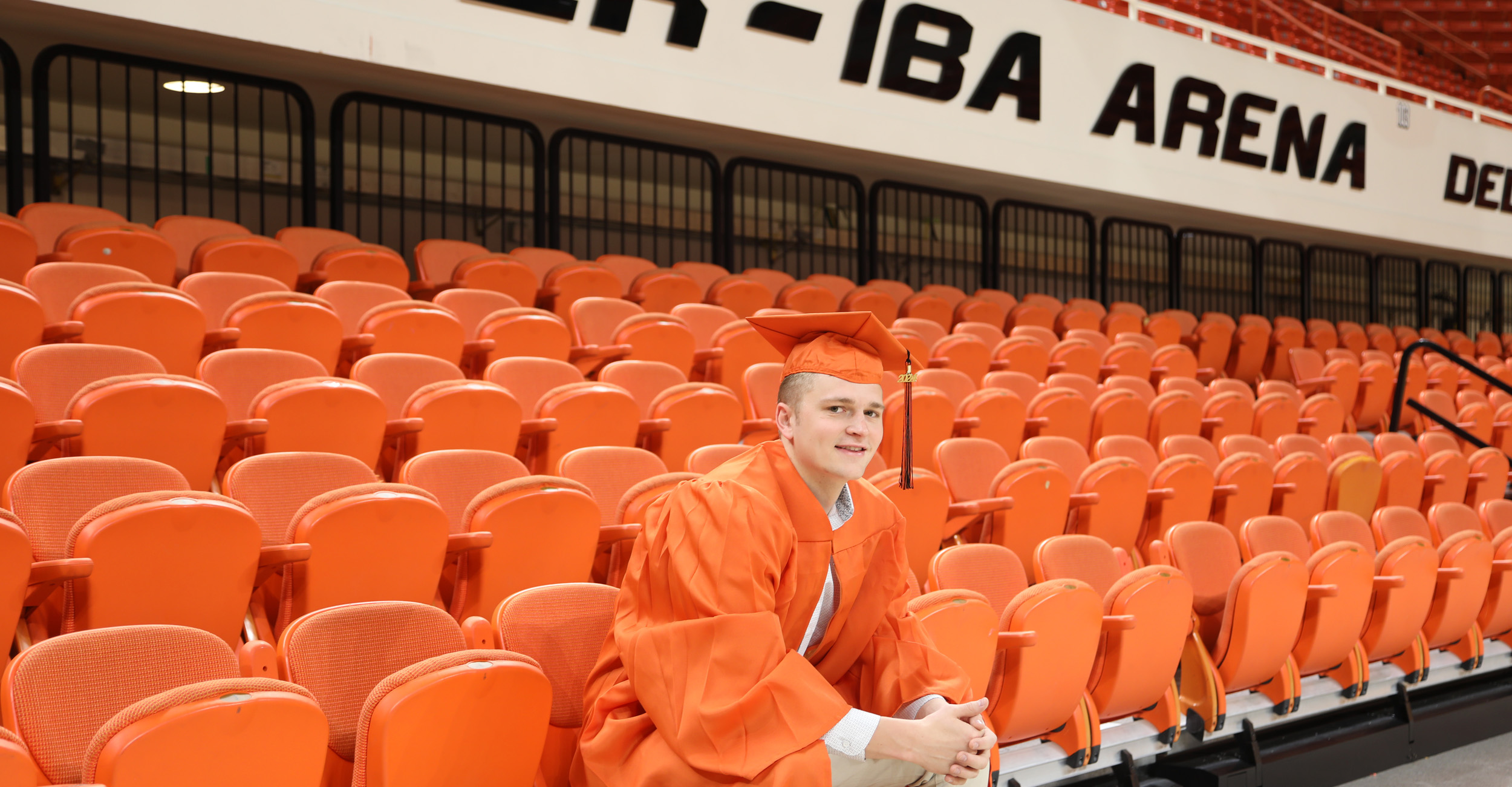 A Caucasian man wears an orange cap and gown while sitting in orange chairs in a basketball arena.