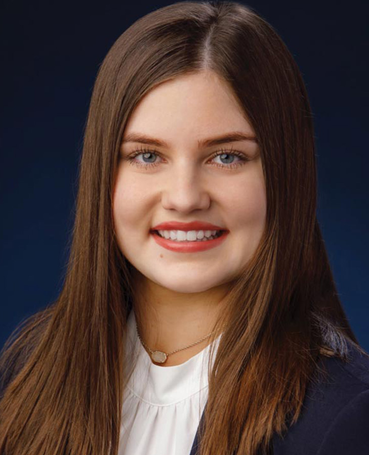 A young woman with long brown hair wearing a white shirt, smiling softly against a neutral background