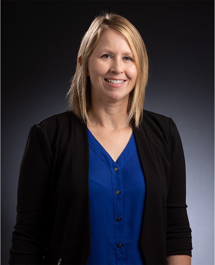 A woman wearing a blue shirt and a black jacket stands confidently, showcasing a professional appearance.